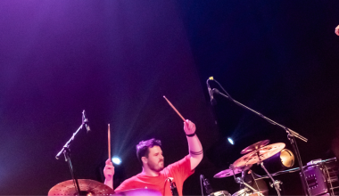 A picture featuring a man holding a microphone while dancing, and a woman dancing. Her arm is outside the picture frame, but it is likely that she is also holding a microphone. They are flanked by a drummer and a keyboard player. The image is of the band Sauwestari in concert.