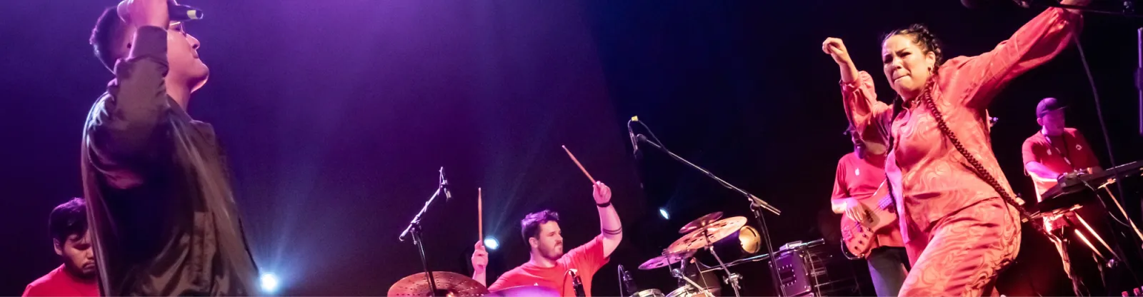 A picture featuring a man holding a microphone while dancing, and a woman dancing. Her arm is outside the picture frame, but it is likely that she is also holding a microphone. They are flanked by a drummer and a keyboard player. The image is of the band Sauwestari in concert.